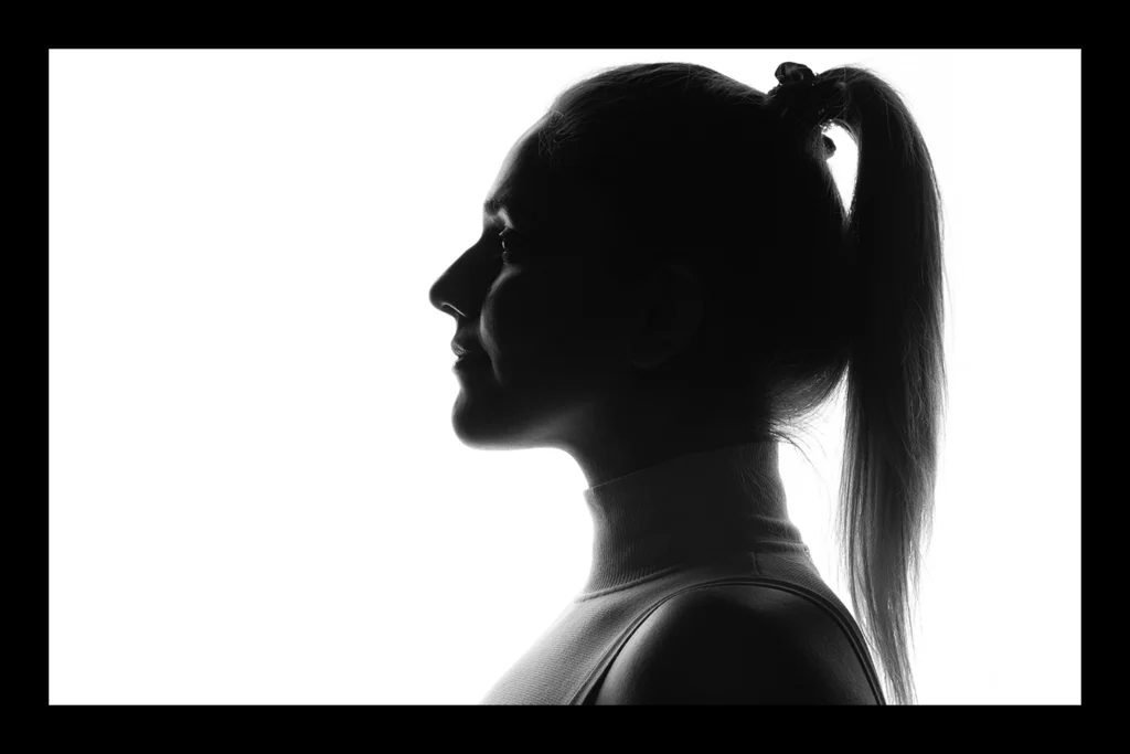 Das Bild zeigt eine Frau im Profil von Schulter bis Kopf, trägt ein weißes Oberteil mit Rollkragen und ohne Ärmel. Die Langen Haare sind zu einem Zopf gebunden. Durch das weiße Gegenlicht vom Hintergrund ergibt sich eine Silhouette. Das Bild ist in schwarz weiß
